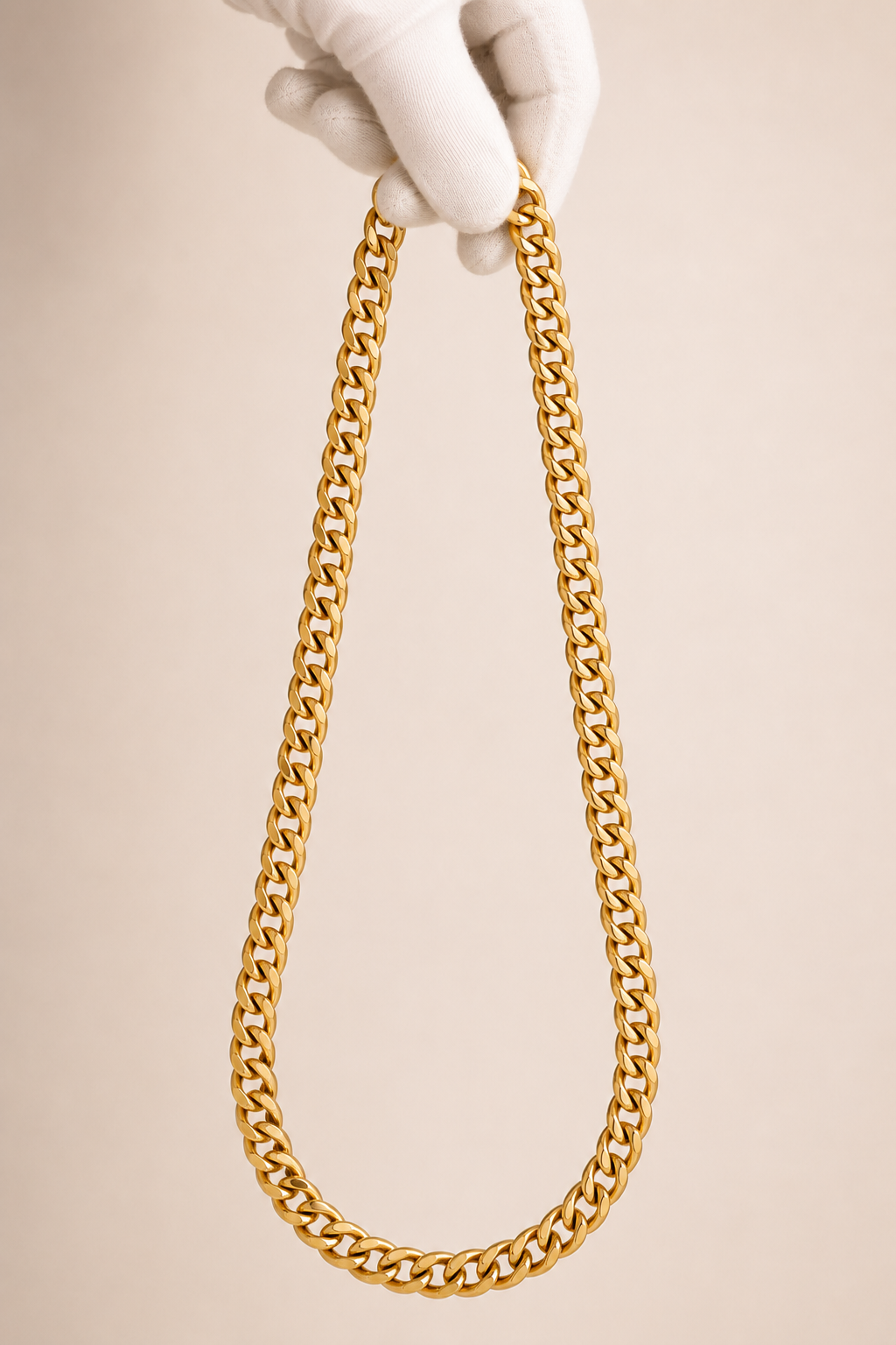A gold-toned chunky curb link chain necklace held up by a white-gloved hand against a plain warm beige background.