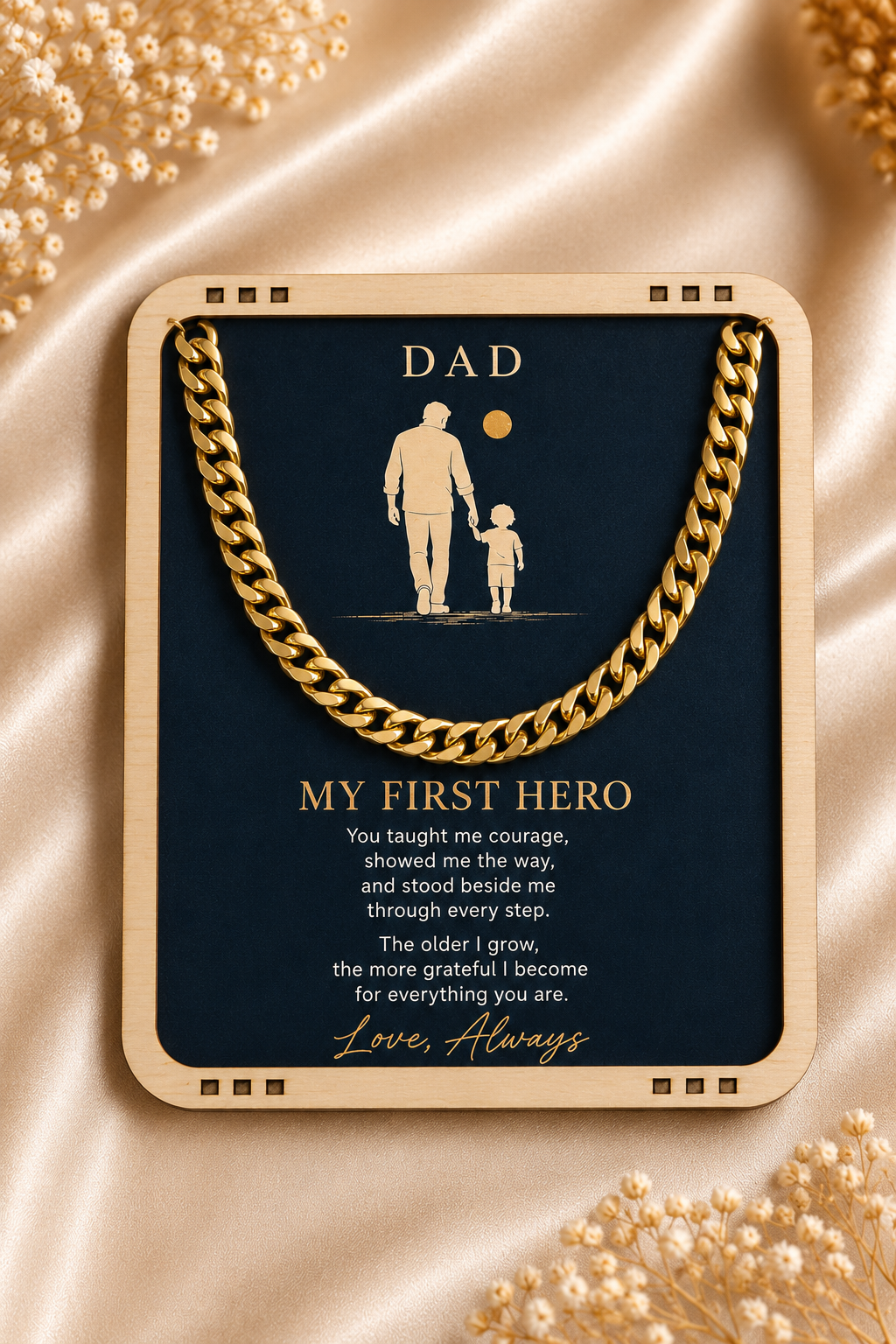 A framed gold-toned necklace with a dark navy card underneath that reads 'DAD' and 'MY FIRST HERO' with a sentimental message. The necklace and card are centered on a champagne satin fabric with decorative gold beads in the corners.