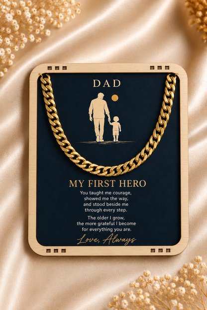 A framed gold-toned necklace with a dark navy card underneath that reads 'DAD' and 'MY FIRST HERO' with a sentimental message. The necklace and card are centered on a champagne satin fabric with decorative gold beads in the corners.