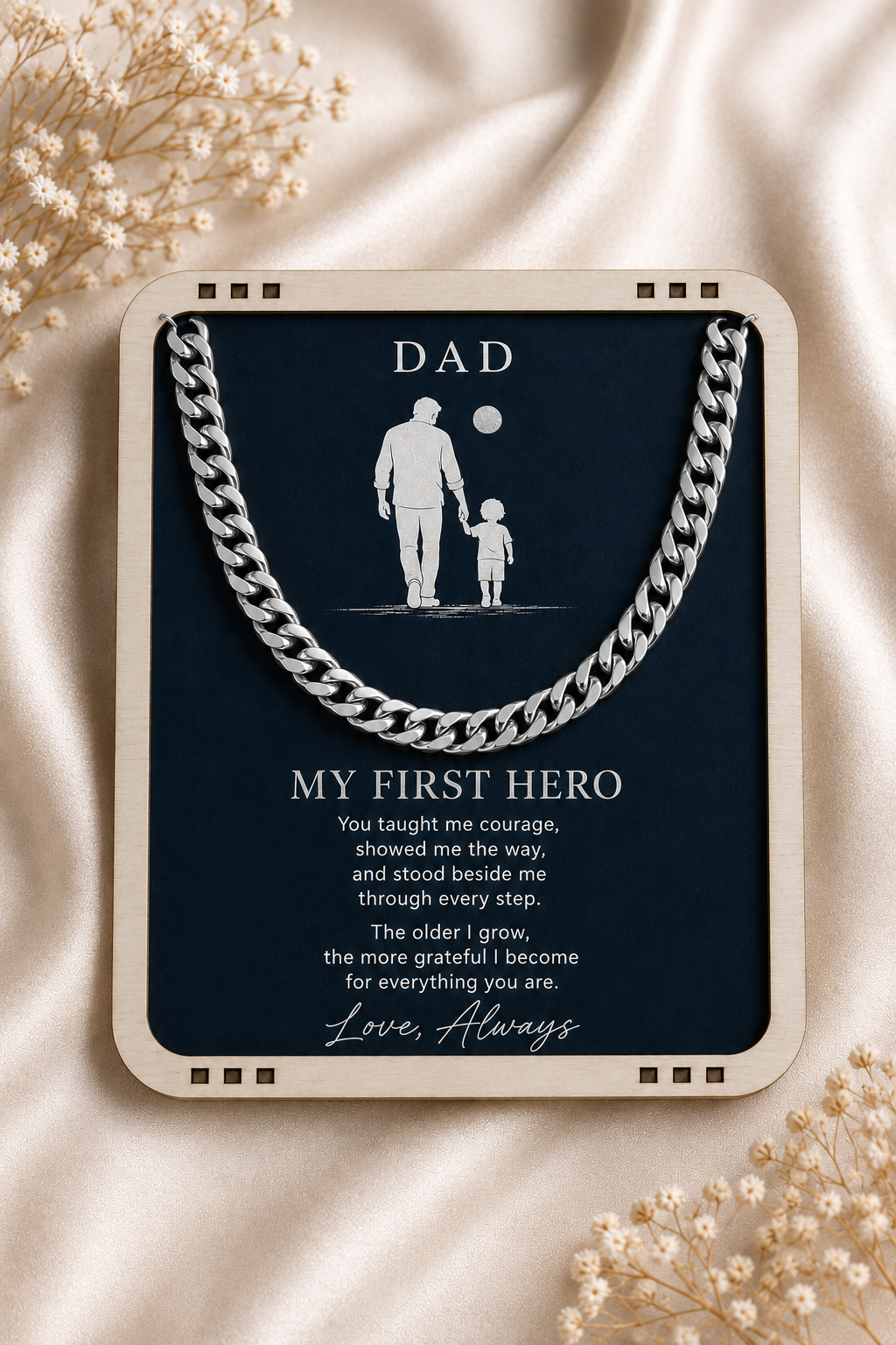 A framed memorial-style plaque or gift card reading 'DAD' and 'MY FIRST HERO' with a silver chain illustration across the top, placed on draped cream fabric with small dried floral props around it.