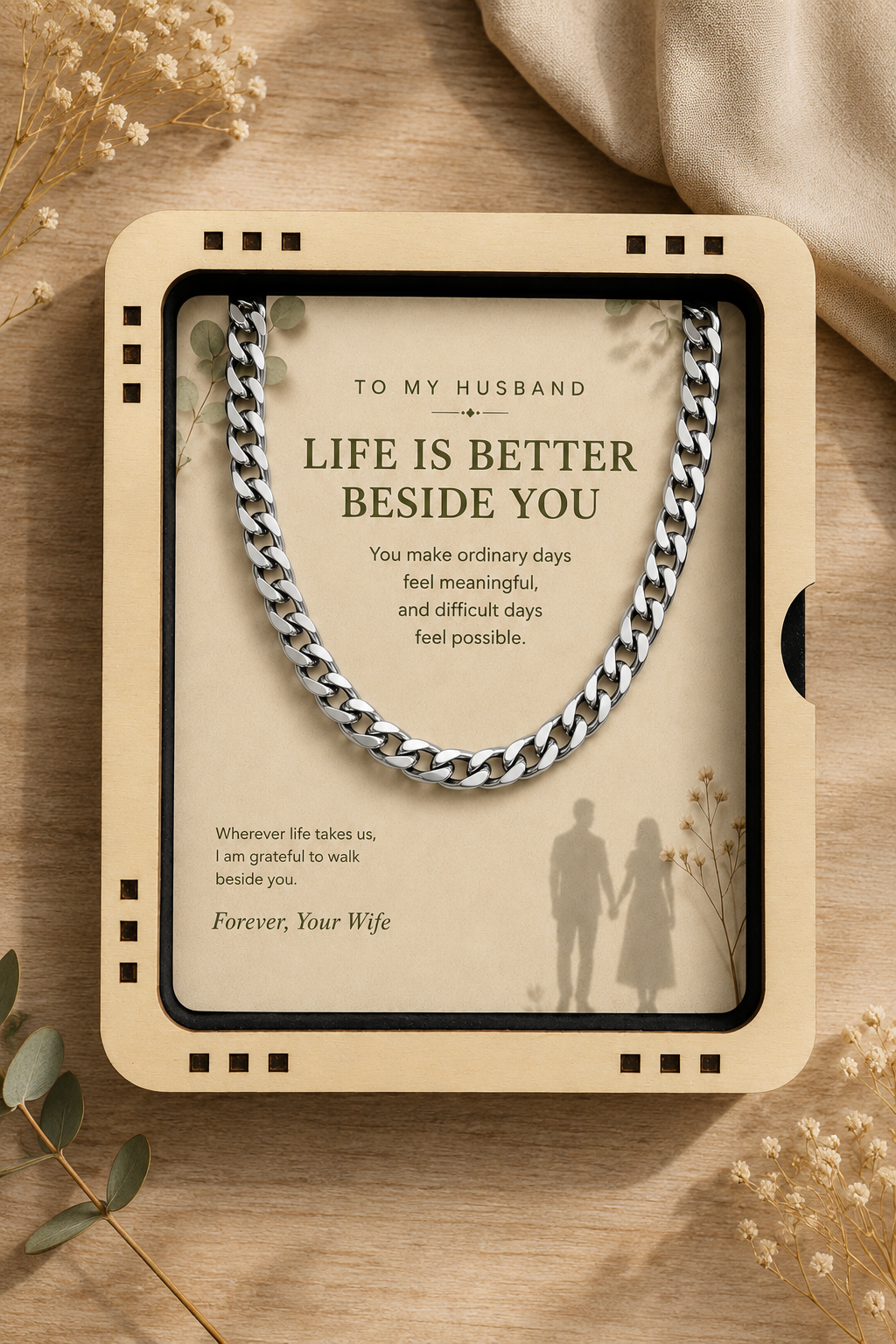A framed sentimental gift box or card on a wooden surface, featuring a silver chain necklace and a printed message for a husband: 'Life is better beside you' with a silhouette of a couple and the text 'Forever, Your Wife.'.