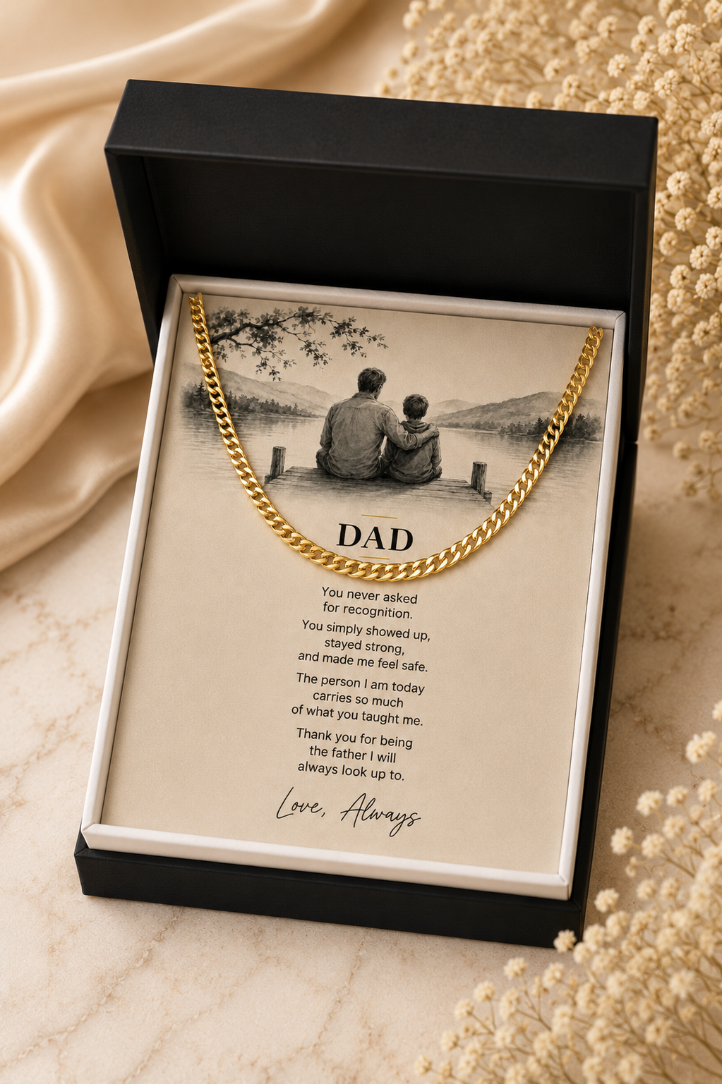 A gold-colored chain necklace presented inside an open black gift box. The necklace is displayed over a printed message card addressed 'DAD' with a sepia-toned photo of two people sitting by a lake.