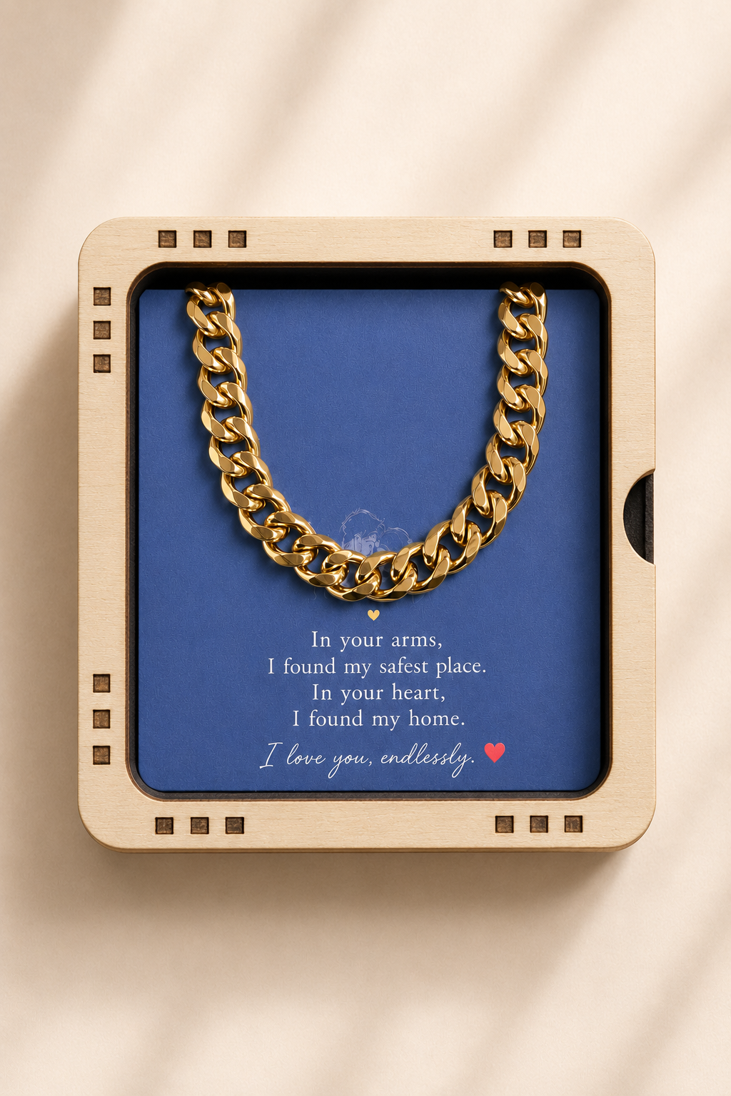 A gold Cuban link chain necklace presented inside a square beige gift box with a blue insert card featuring a romantic message, photographed from above on a light cream surface with soft shadow overlays.