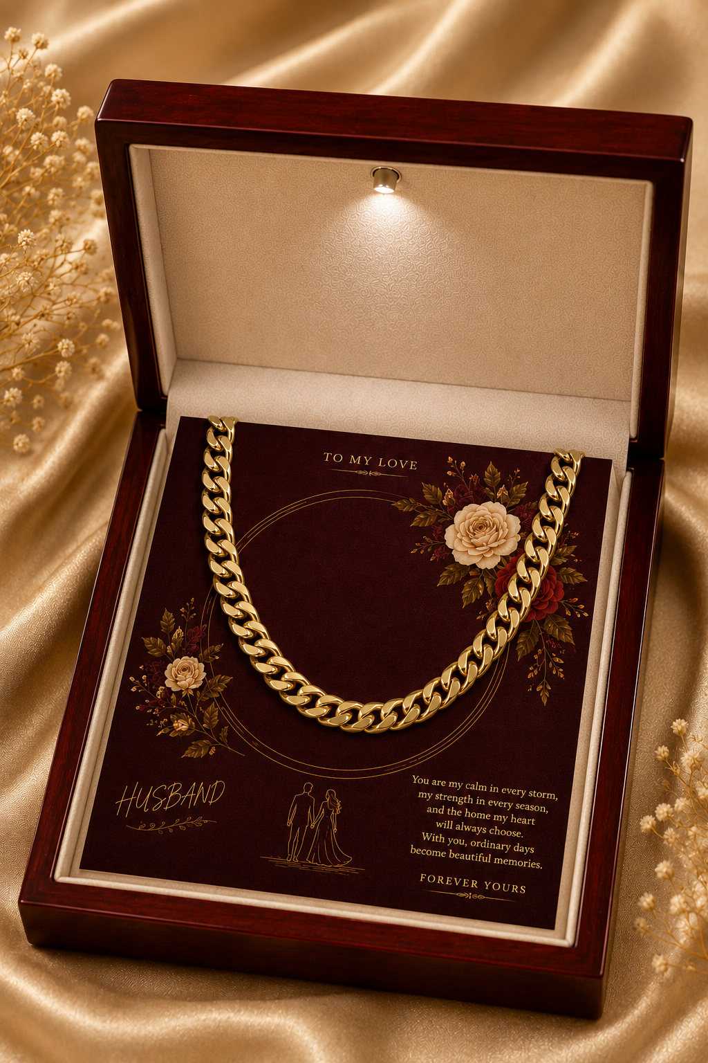 A gold Cuban link chain necklace presented inside an open dark wood gift box with a cream velvet interior and a printed message card. The box is photographed on a champagne satin fabric background with decorative pearl strands around it.
