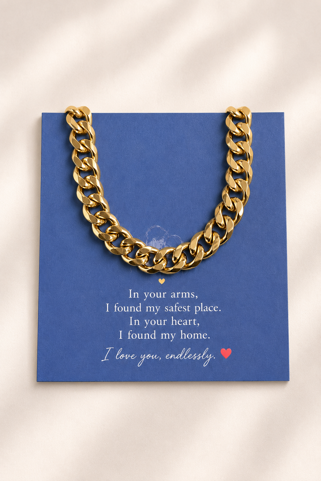 A gold chain necklace draped over a blue card with white sentimental text and a small red heart, photographed on a softly lit cream background.