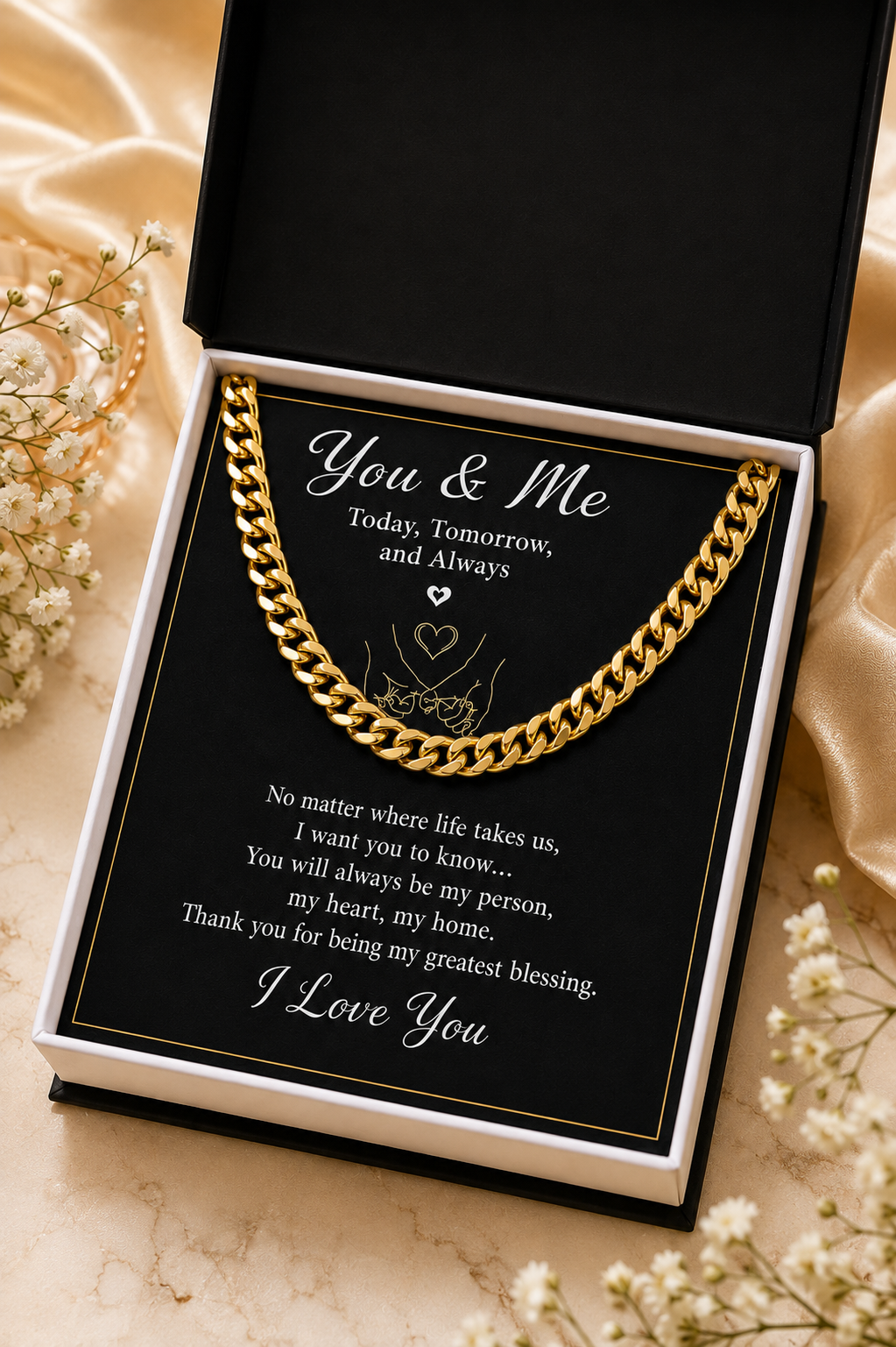 A gold chain necklace presented inside an open black gift box with a printed message card. The necklace is draped across the black insert, and the box is styled with satin fabric and small white flowers on a light beige surface.