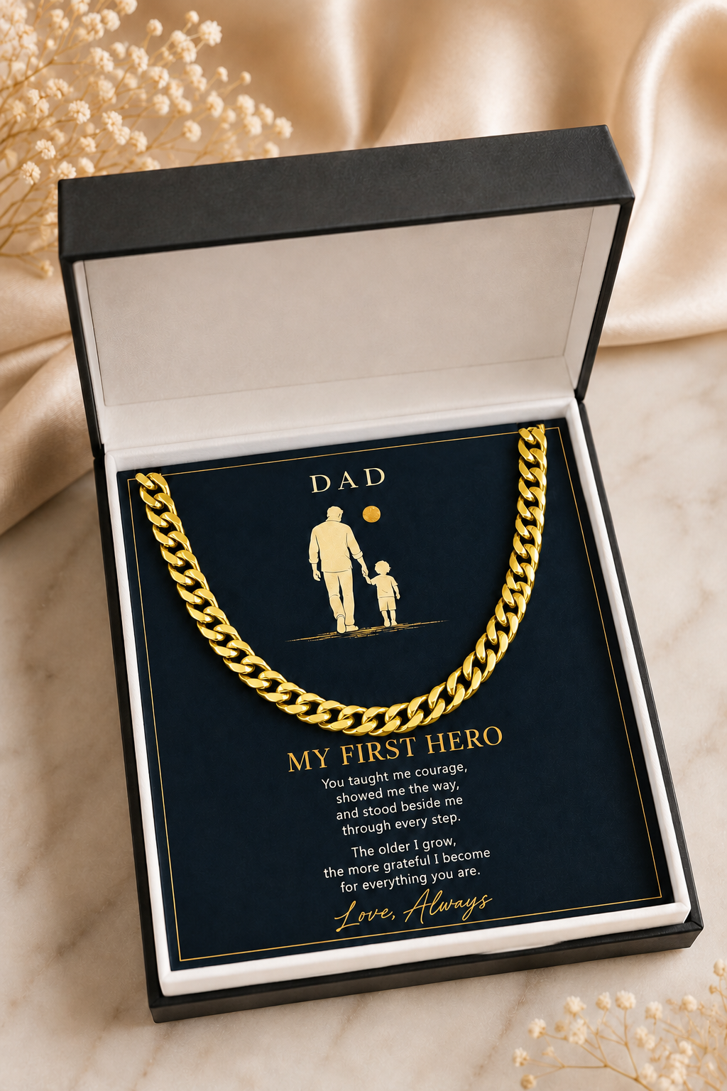 A gold chain necklace presented inside an open black gift box with a navy card that reads 'DAD MY FIRST HERO' and includes a small father-and-child silhouette. The box is styled on a light marble surface with soft beige fabric and dried flowers in the background.