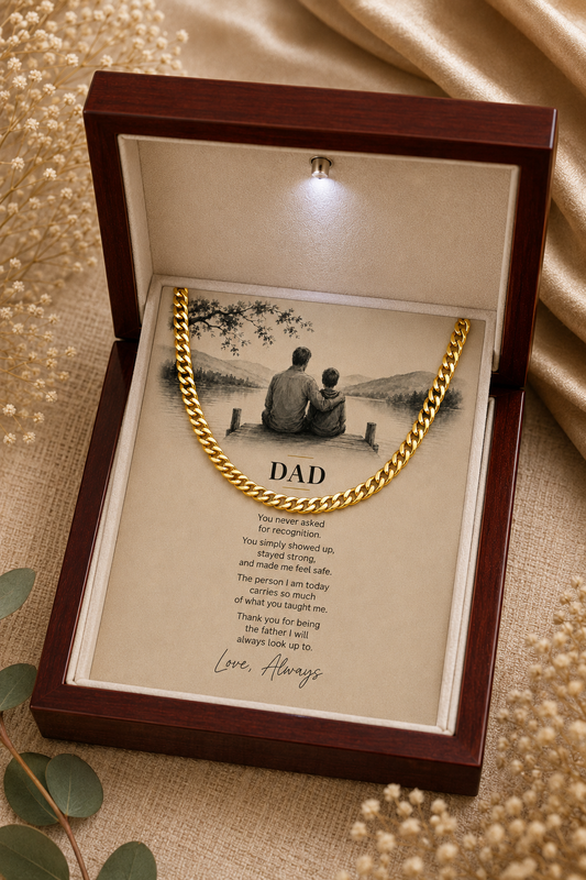 A gold chain necklace presented inside an open dark wood gift box with a printed message card and photo of two people sitting by a lake. The box is photographed on a beige fabric surface with dried flowers and green leaves around it.