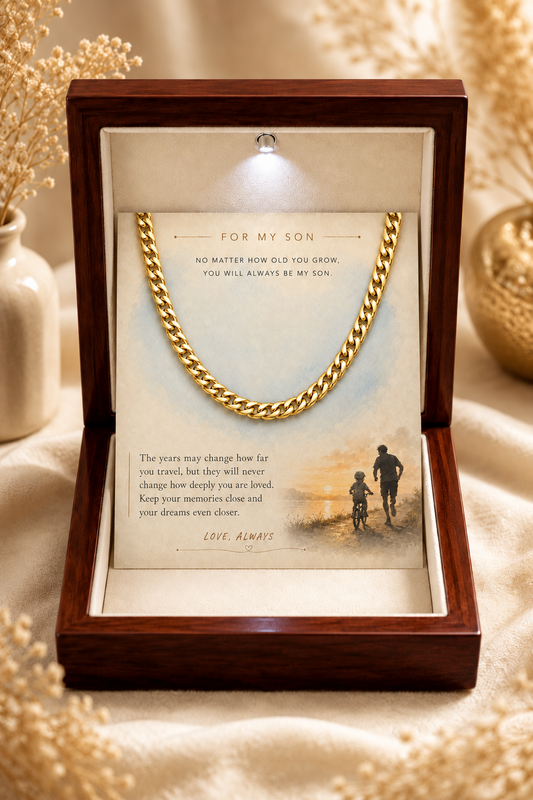 A gold chain necklace presented inside an open wooden gift box with a printed sentimental message card that reads 'FOR MY SON'. The box is set on a beige fabric surface with dried flowers and warm decorative props in the background.