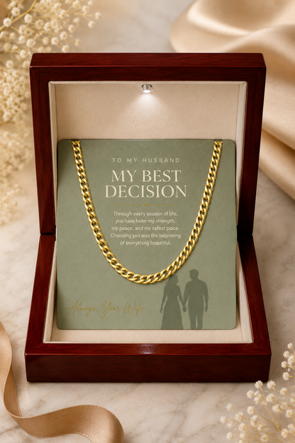 A gold curb chain necklace presented inside an open dark wood gift box with a cream insert. The chain is draped over a sage green card that reads 'TO MY HUSBAND MY BEST DECISION' with a short message and a silhouette of a couple.