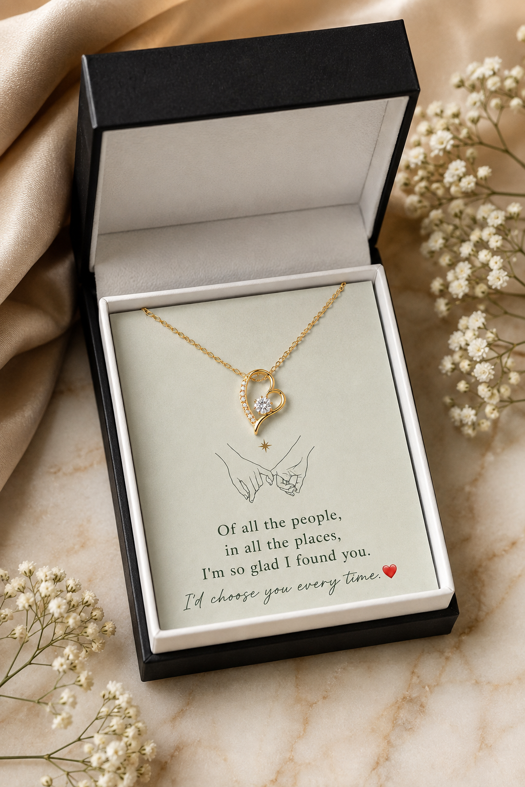 A gold heart-shaped necklace with a clear crystal center, presented in an open black gift box with a printed message card. The box is styled on a marble surface with beige fabric and dried baby's breath flowers.