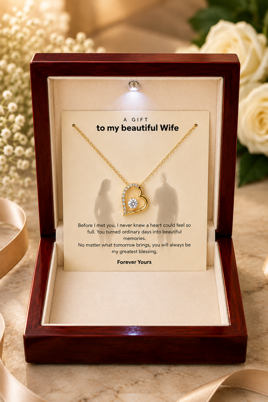 A gold heart-shaped pendant necklace with a clear central gemstone, presented inside an open dark wood gift box with a printed message card addressed 'to my beautiful Wife'.