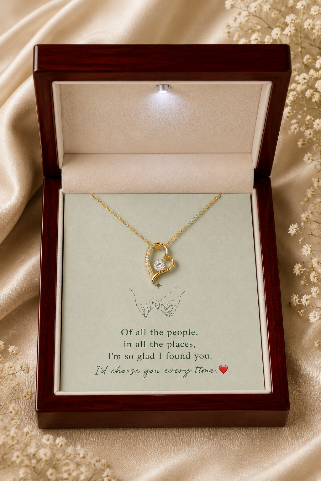 A gold heart-shaped pendant necklace with a clear central stone, presented in an open dark wood gift box with a cream insert card. The box sits on draped champagne satin fabric with small white flowers around it.