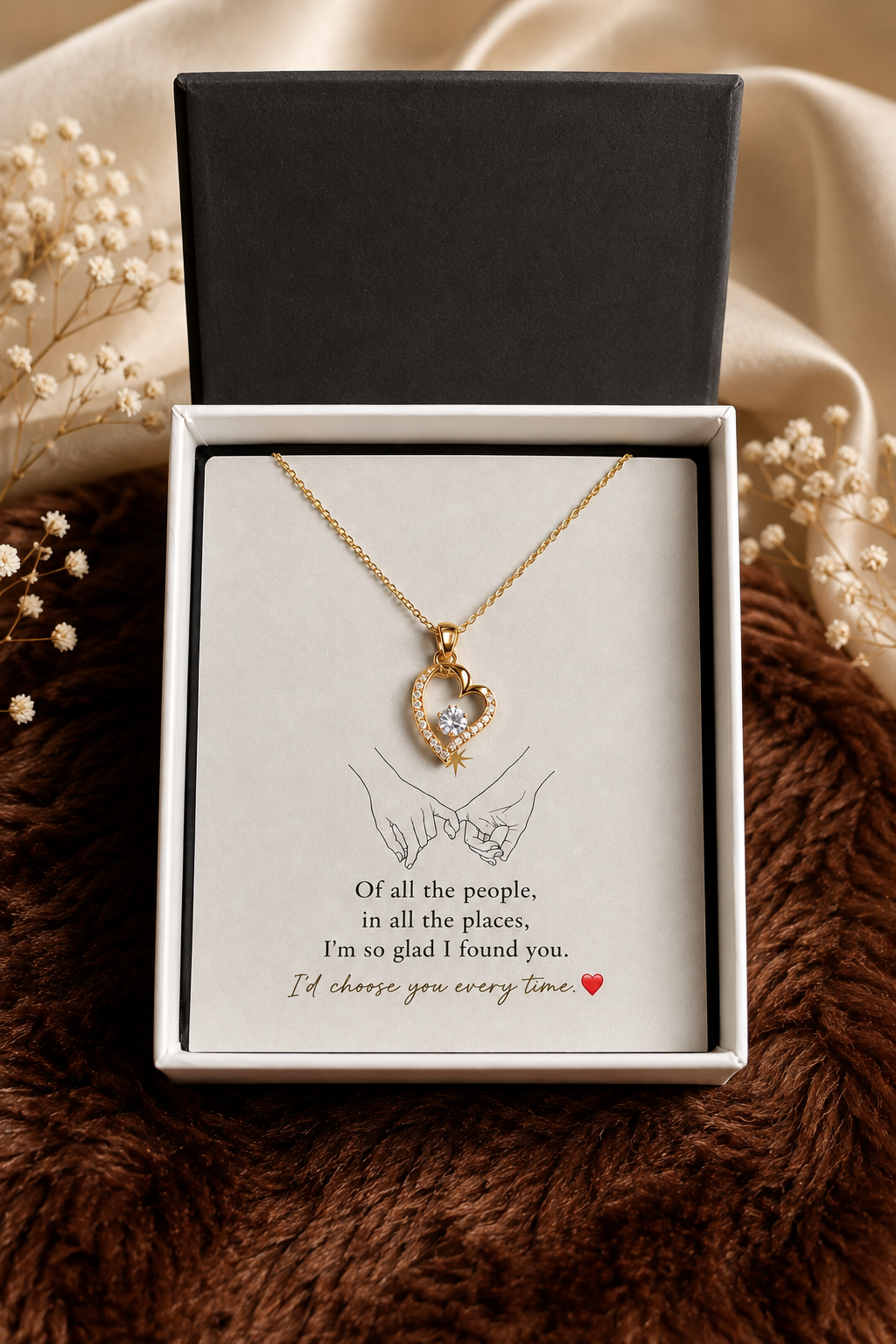 A gold heart-shaped pendant necklace with a clear heart in the center, displayed inside an open white gift box with a black lid. The necklace is presented on a printed card with a line drawing of hands and a sentimental quote.