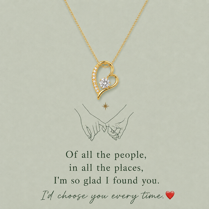 A gold heart-shaped pendant necklace with a small clear gemstone, displayed on a light sage green background with romantic text and a simple illustration of two hands holding.