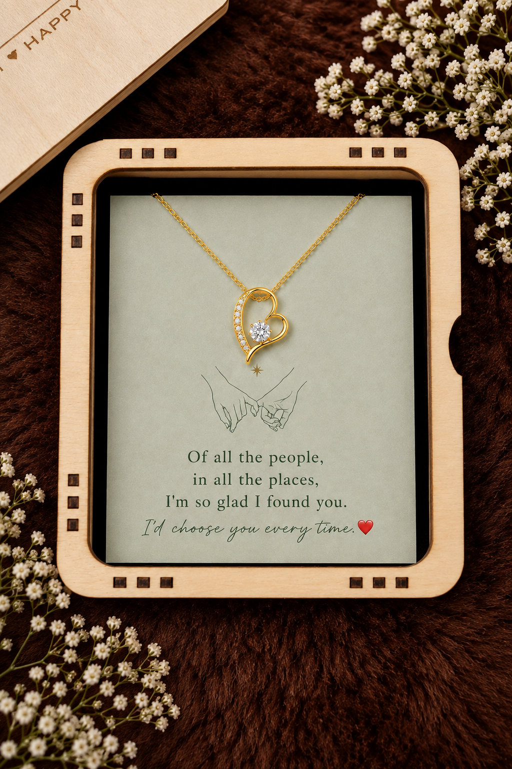 A gold heart-shaped pendant necklace with a small clear stone is displayed on a printed card inside a light wood frame, styled with dried flowers on a dark brown textured surface.