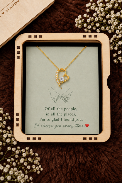 A gold heart-shaped pendant necklace with a small clear stone is displayed on a printed card inside a light wood frame, styled with dried flowers on a dark brown textured surface.