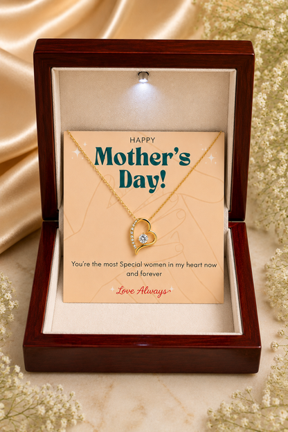 A gold heart necklace with a small clear gemstone is displayed inside an open dark wood gift box. The necklace is attached to a beige card that reads 'Happy Mother's Day!' with a warm spotlight from above.