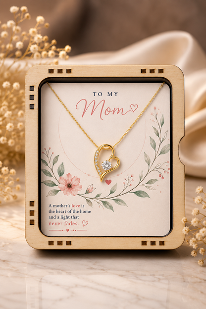 A gold heart pendant necklace displayed on a decorative card inside a square wooden gift box, with floral artwork and the message 'TO MY Mom' on a warm beige background.
