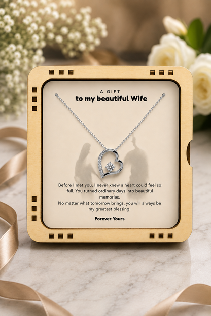 A heart-shaped silver necklace with a small clear gemstone pendant, presented on a cream card inside a gold-toned square gift box. The card reads 'A GIFT to my beautiful Wife' with a romantic message below, set against a softly blurred floral background with beige ribbons.