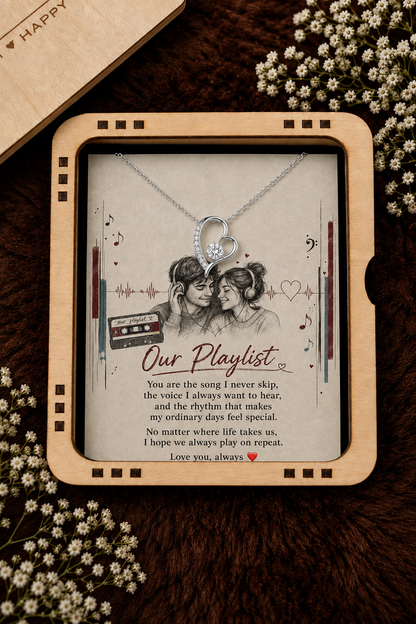 A heart-shaped silver pendant necklace displayed inside a square wooden gift box with a sentimental 'Our Playlist' message, surrounded by small white flowers on a dark brown textured surface.