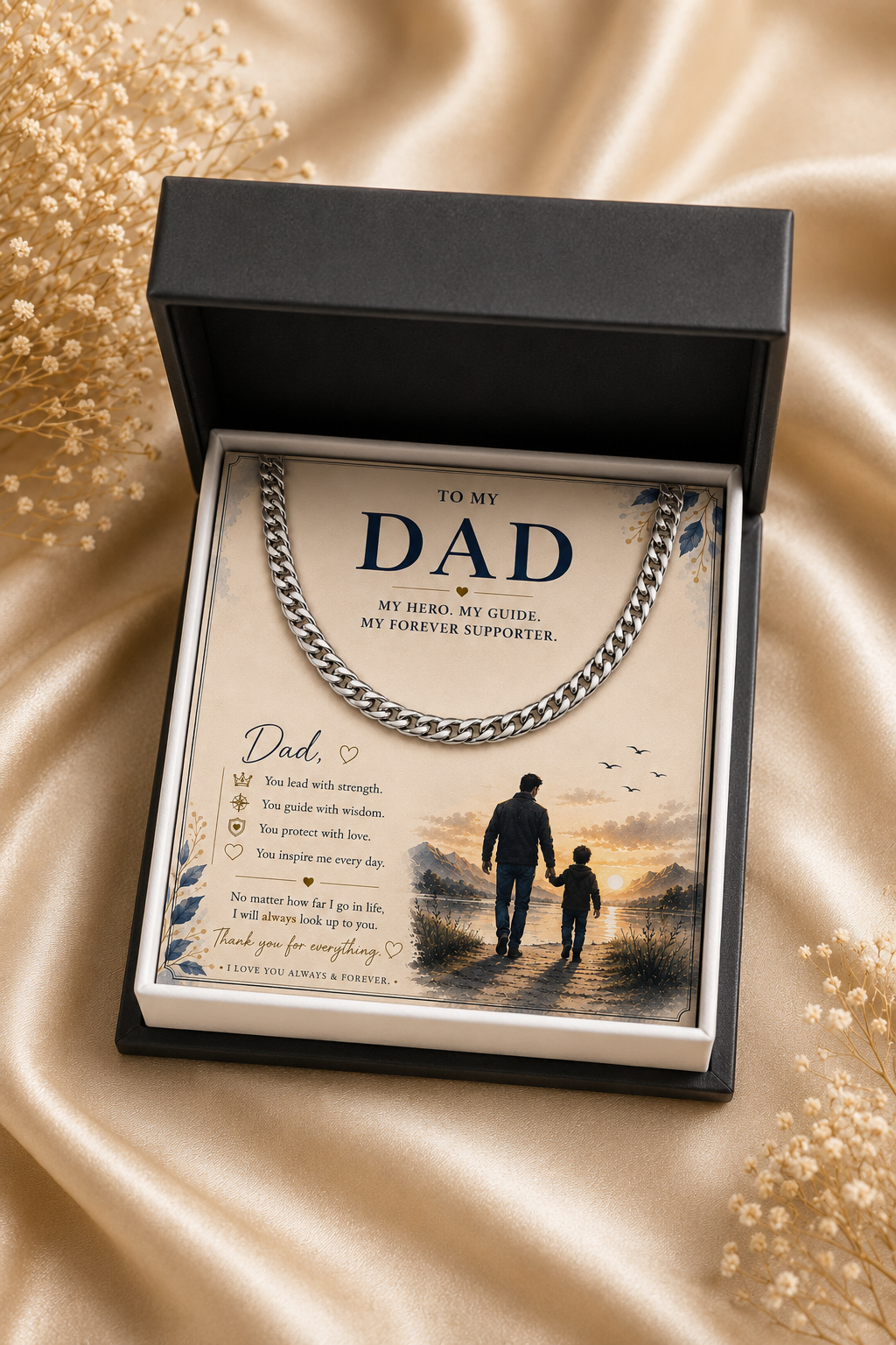 A silver-toned chain necklace presented inside an open black gift box with a printed message card addressed 'To My Dad.' The card includes a family silhouette illustration and sentimental text, set on a beige satin fabric background with decorative dried flowers.