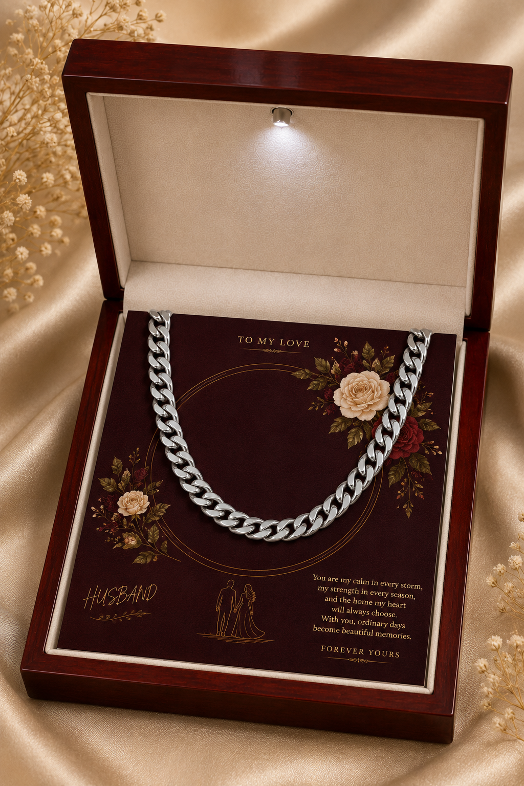 A silver-toned chain necklace presented inside an open dark wood gift box with a cream interior and printed message card. The box is photographed on a beige satin fabric background with decorative dried flowers around it.