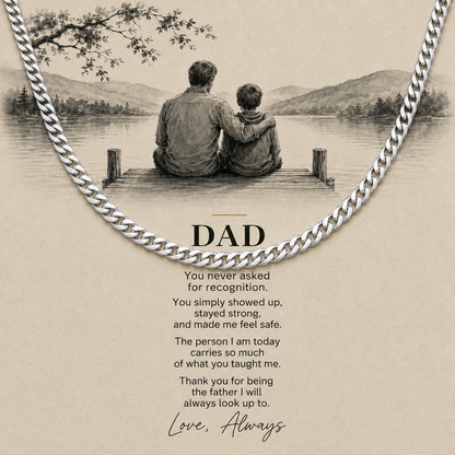 A silver chain necklace draped over a beige printed card featuring a black-and-white illustration of a father and child sitting on a dock by a lake, with the word “DAD” and a heartfelt message below.
