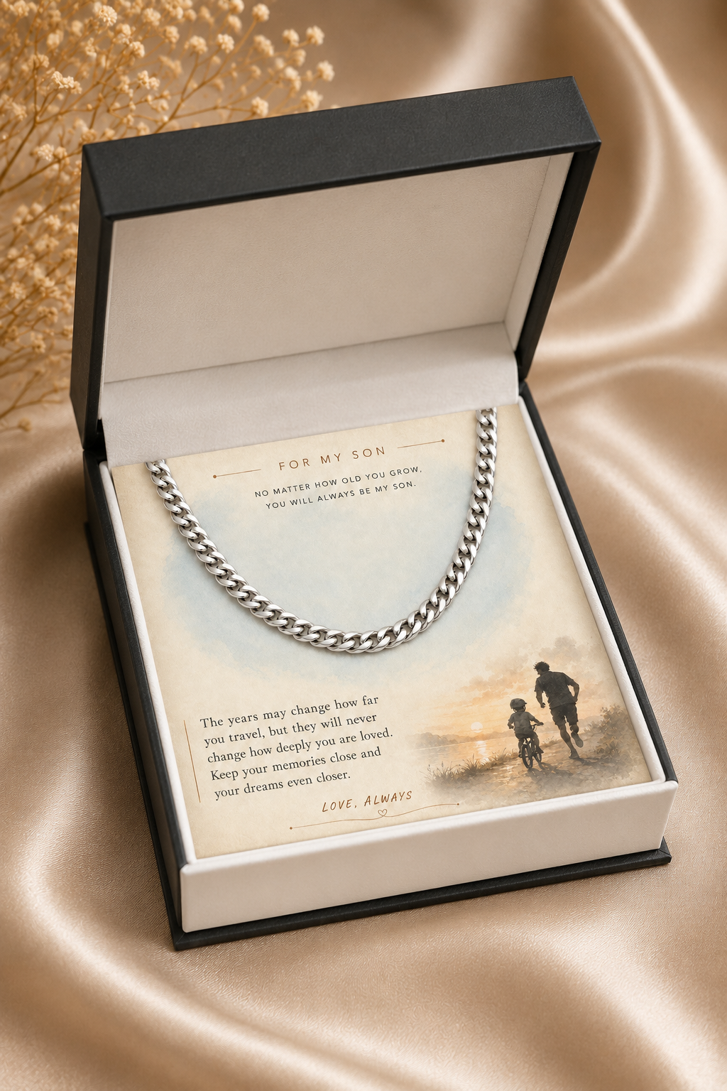 A silver chain necklace presented in an open black gift box with a cream insert card that reads 'FOR MY SON' and includes a family silhouette. The box is photographed on a warm beige satin fabric background with dried floral decor in the corner.