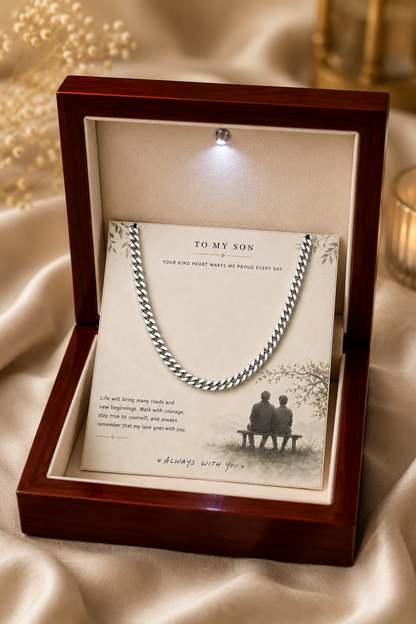 A silver chain necklace presented inside an open dark wood gift box with a cream insert card addressed 'TO MY SON' and a small light bulb under the lid, set on draped satin fabric.