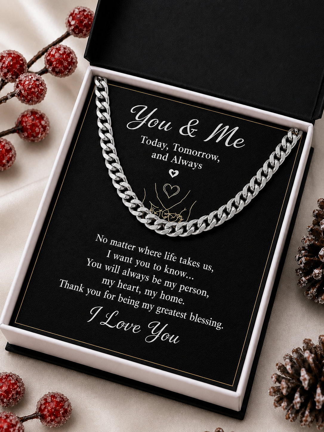 A silver chain necklace presented inside an open black gift box with a printed message card reading 'You & Me' and a sentimental poem. The box is styled on a light fabric surface with red berries and pinecones around it.