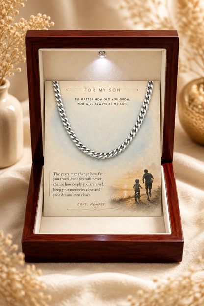 A silver chain necklace presented inside an open dark wood gift box with a cream insert. The insert includes a printed message for a son and a small illustration of a parent and child walking by the sea.