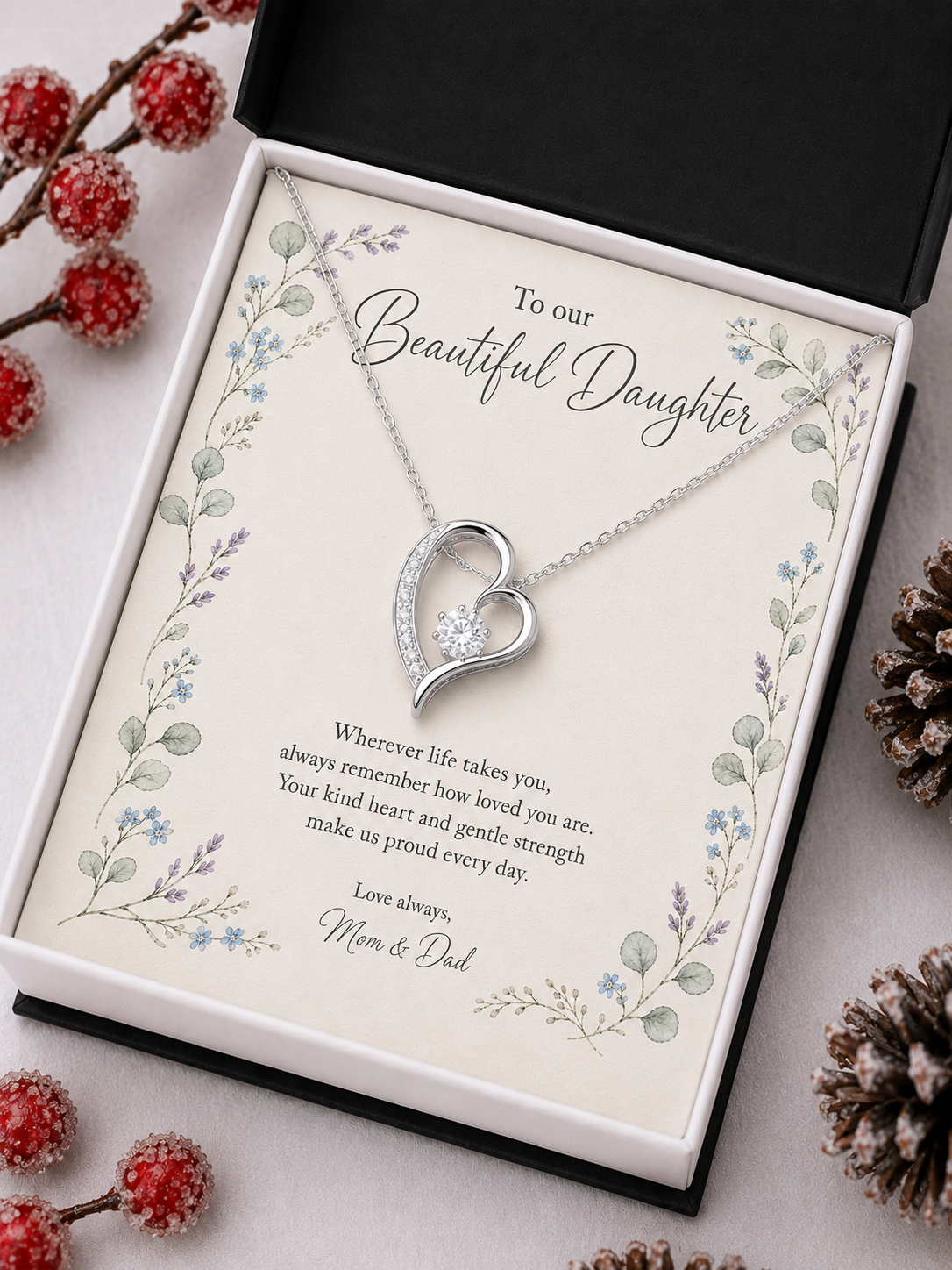 A silver heart-shaped pendant necklace displayed on a printed gift card inside an open black presentation box. The card reads 'To our Beautiful Daughter' and includes a floral border and a message for Mom & Dad.