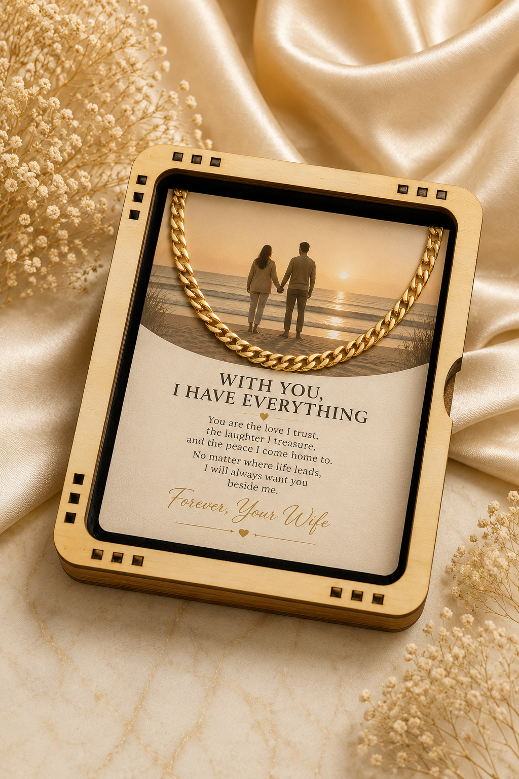 A square gold-toned keepsake box with a hinged lid, shown open on a beige marble-like surface with draped satin fabric and dried flowers. The inside features a printed message for a wife and a photo of a couple standing by a beach at sunset, with a gold chain draped across the top.
