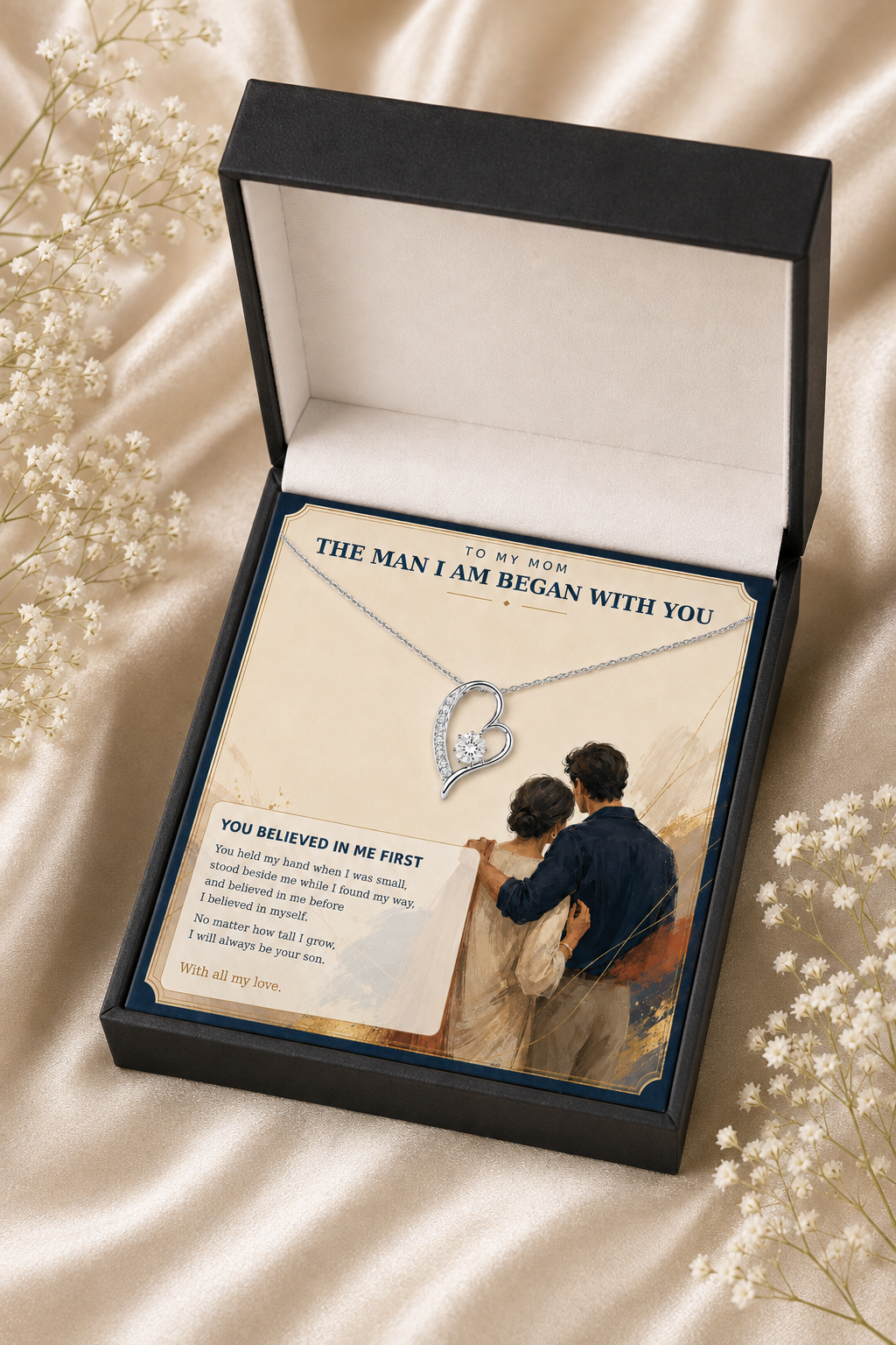 An open black gift box containing a heart-shaped silver pendant necklace displayed on a printed card with sentimental text and an illustration. The box is photographed on a cream satin fabric with small white flowers around it.