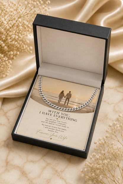 An open black gift box containing a silver chain necklace and a printed message card, styled on a beige marble surface with satin fabric and dried flowers around it.