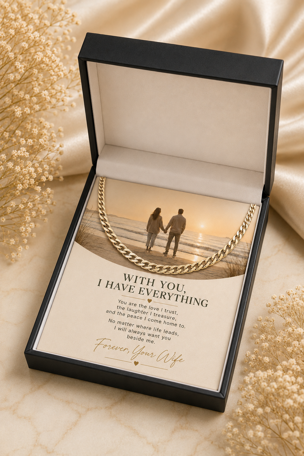 An open black gift box with a printed message card inside, featuring a gold chain necklace draped over the lid. The card reads 'WITH YOU, I HAVE EVERYTHING' and includes a photo of a couple on a beach.