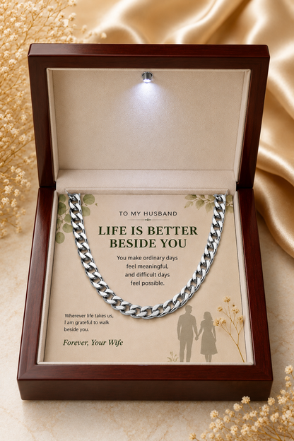 An open dark wood gift box with a built-in light, displaying a silver chain necklace over a printed message card addressed to a husband. The box is set on a beige satin fabric background with dried floral decorations.