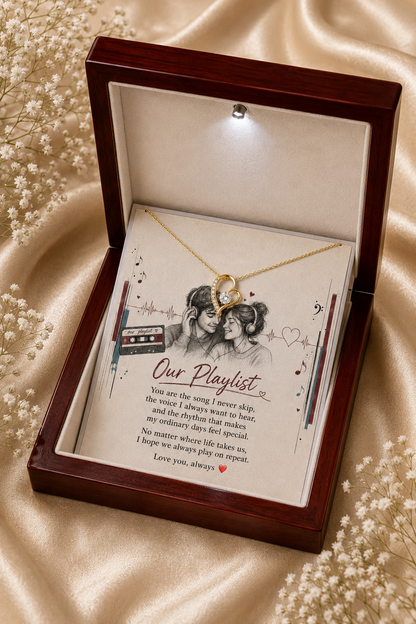 An open dark wood keepsake box on beige satin fabric, displaying a gold heart necklace over a printed card that reads 'Our Playlist' with a black-and-white illustration of two girls.
