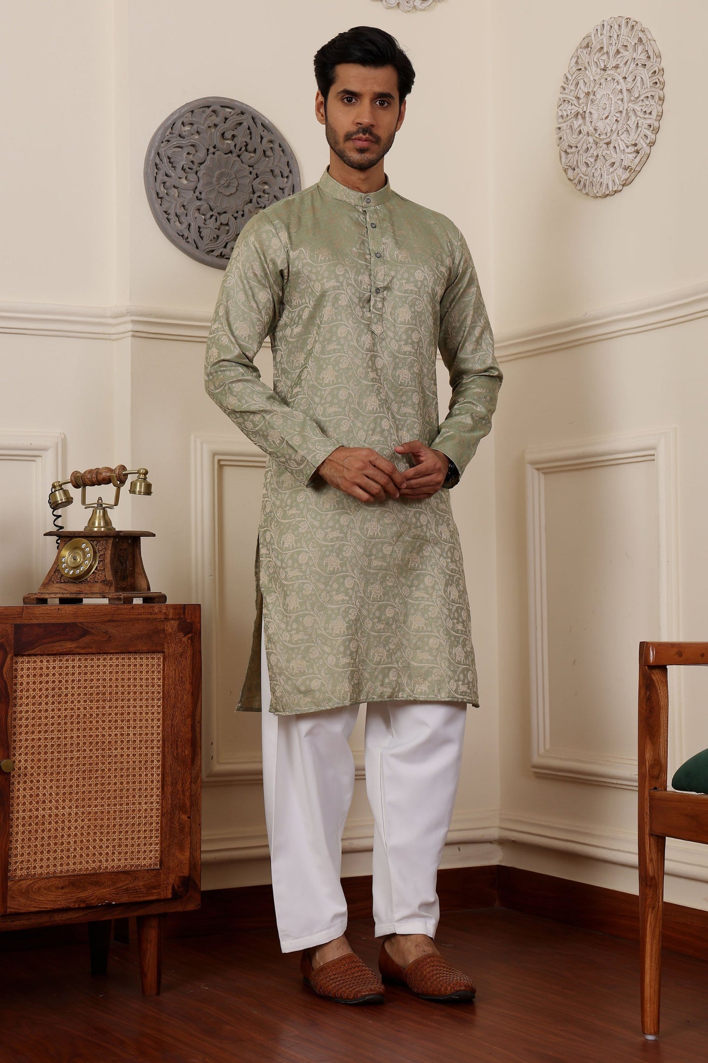 A man standing in an elegant olive green kurta with Wevon jacquard work, paired with white trousers and a dupatta, in a room with traditional decor.