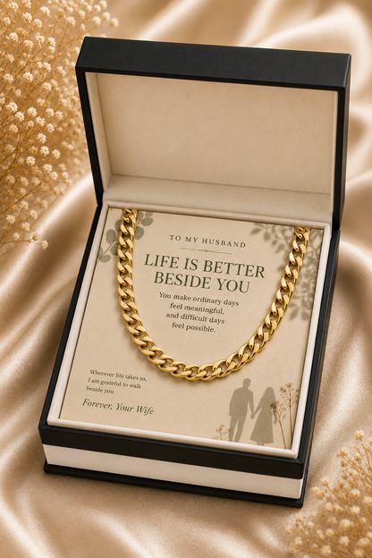 A gold chain necklace presented in an open black gift box with a cream insert card that reads 'TO MY HUSBAND LIFE IS BETTER BESIDE YOU' and 'Forever Your Wife.' The box is photographed on a satin-like beige surface with decorative dried flowers around it.