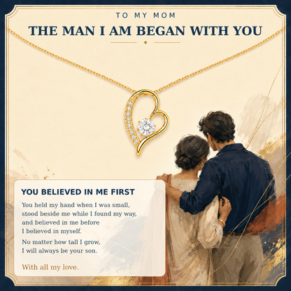 A sentimental gift card or print featuring a gold heart-shaped pendant necklace with a central clear gemstone, shown above an illustration of a father and son. The card has a cream background with navy blue text reading 'TO MY MOM THE MAN I AM BEGAN WITH YOU' and a poem below.