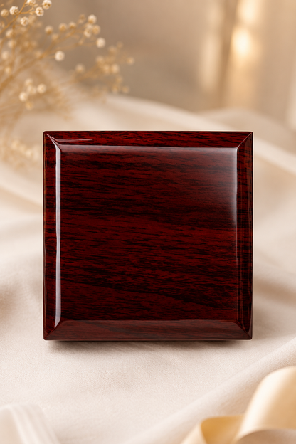 A square glossy dark reddish-brown wooden box with a polished finish, set against a soft beige fabric backdrop with blurred dried flowers.