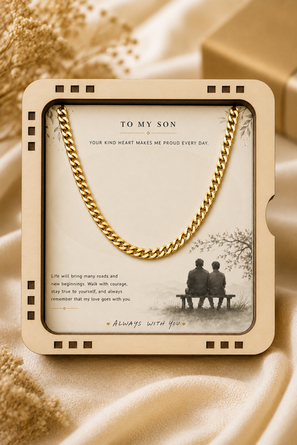 A gold chain necklace presented inside a square cream-colored gift box with a sentimental message for a son, set against a draped beige fabric background.