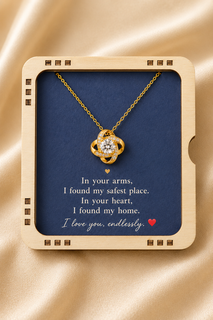 A gold necklace with a heart-shaped pendant displayed inside a square cream-colored gift box with a navy blue insert. The insert features a romantic quote printed in white text with a small red heart at the bottom.