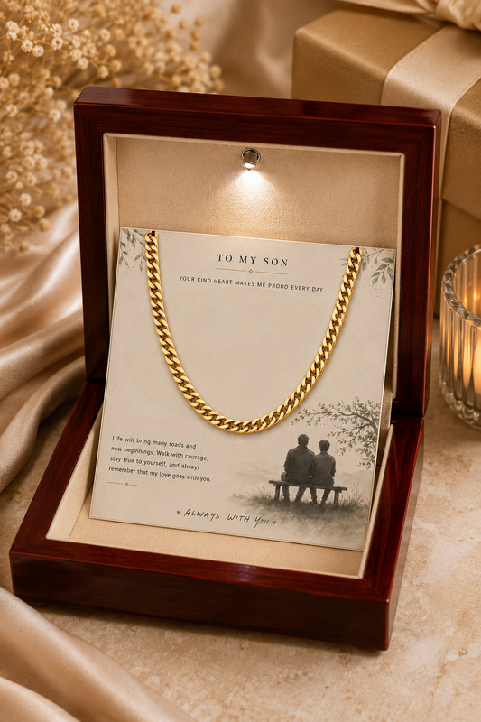 A gold chain necklace displayed inside an open dark wood gift box with a cream insert card addressed 'TO MY SON.' The necklace is draped over the card, and the box sits on a satin fabric surface with a candle and wrapped gift in the background.