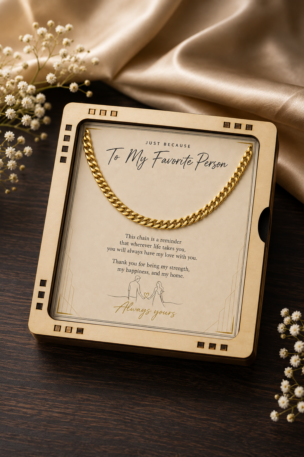 A gold chain necklace presented in a square cream-colored gift box with a transparent window, placed on a dark wooden surface with satin fabric and small white flowers around it. The box has a printed message and the chain is draped across the top.