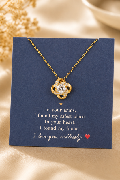 A gold necklace with a clear heart-shaped gemstone pendant, displayed on a navy blue gift card with a sentimental quote. The card is set against a soft beige satin background with dried flowers.