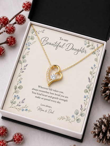 A gold heart pendant necklace with a small clear stone is displayed on a printed gift card inside an open black and white presentation box. The card reads 'To our Beautiful Daughter' and includes floral border artwork and a message for 'Mom & Dad.