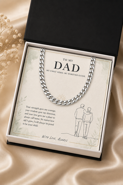 A silver chain necklace presented in a black gift box with a cream message card addressed 'TO MY DAD.' The necklace is draped across the card, and the box sits on a satin fabric surface with dried flowers beside it.